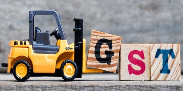 Yellow plastic forklift hold letter G to complete word GST (Abbreviation of Goods and Services Tax) on wood background